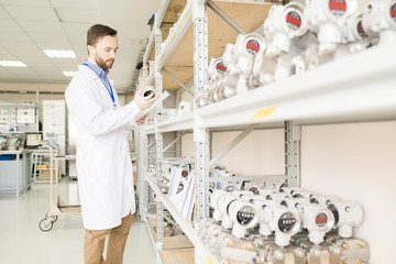 Serious concentrated bearded quality control engineer in lab coat reading document and examining number of measuring device in transducer factory warehouse