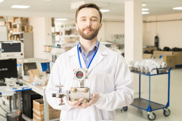Fototapeta na wymiar Smiling confident handsome male specialist of manometer plant holding measuring device and looking at camera in illuminated assembly shop