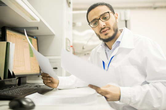 Serious concentrated Arabian male engineer in lab coat and glasses reading papers and examining information while holding expertise in office