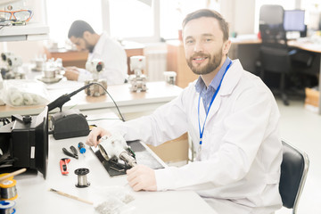 Fototapeta na wymiar Cheerful confident bearded male repairing engineer in lab coat sitting at table with tools and dissembled measuring device and looking at camera
