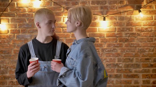 Young interesting boy hipster is standing with cup, his girlfriend is flirting with him, kissing, French kiss, love conception, brick bsckground