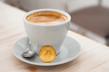 Cryptocurrency golden bitcoin standing on coffee cup. Golden cryptocurrency and white cup hot coffee mug on the soft background.