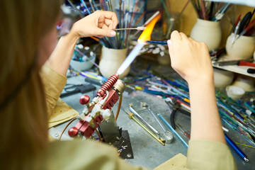 Over the shoulder portrait of young female artist melting glass by gas torch while making beautiful handmade jewelry in glassworking studio