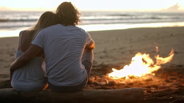 Medium shot with rear view of unrecognizable couple sitting by campfire on beach and looking at ocean