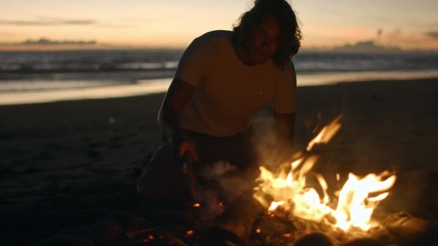 Medium shot of young man with long curly hair tending campfire on beach at dusk