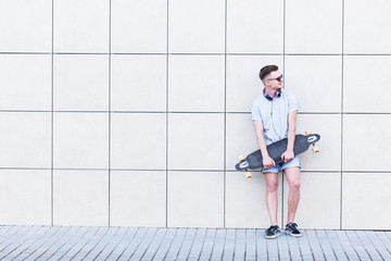 Skateboarder dressed in grey t-shirt and sunglasses holds longboard before modern building bricks wall