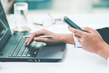 A business woman analyzing investment charts at workplace and holding a smart phone and using laptop in office.