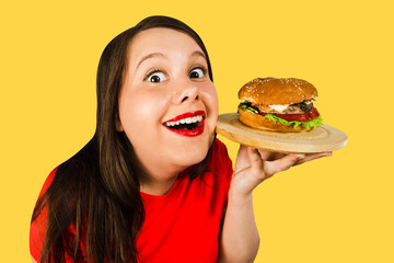 Young smiling fat woman holds burger and smiles isolated on yellow background.