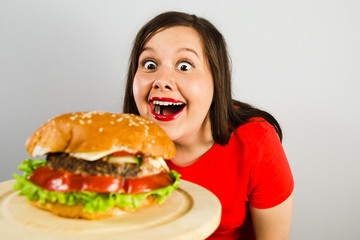 Young fat woman looks surprised at the burger with wide eyes on gray background