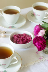 Jam from the petals of the Damascus rose, a cup of green tea and a vase of roses on a light table. Rustic style.