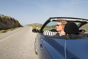 Young blonde woman driving convertible blue rental car without roof on mountain road in Naxos island, Greece