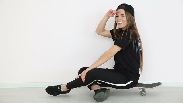 teenage girl in trendy hip hop clothes and cap posing against white wall with skateboard