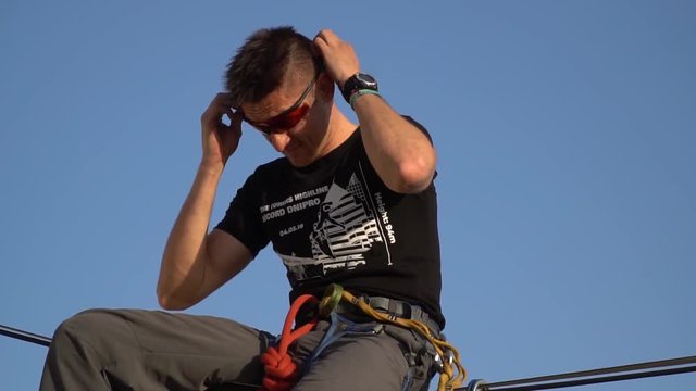 A man on a blue sky background sits on a tight rope and puts on sunglasses