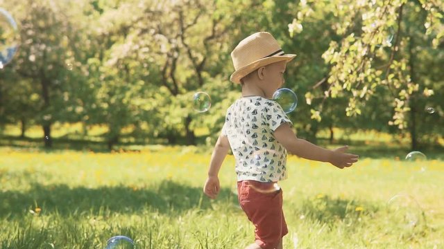 Close up portrait of little boy in hat catching soap bubbles. Little child playing with parents outside on summer sunny day in slow motion