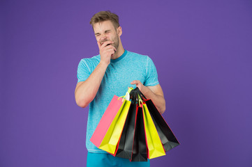 Tired man with shopping bags on violet background. Shopping or sale and cyber monday. Macho with colorful paper bags. Fashion shopper in casual blue tshirt. Holidays preparation and celebration