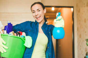 Beautiful young woman in protective gloves is holding a green bucket with equipment for cleaning. Girl looking at camera and smiling