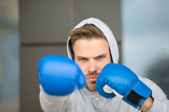 Boxing concept. Man athlete on concentrated face with sport gloves practicing boxing punch, urban background. Boxer with hood on head practices jab punch. Sportsman boxer training with boxing gloves