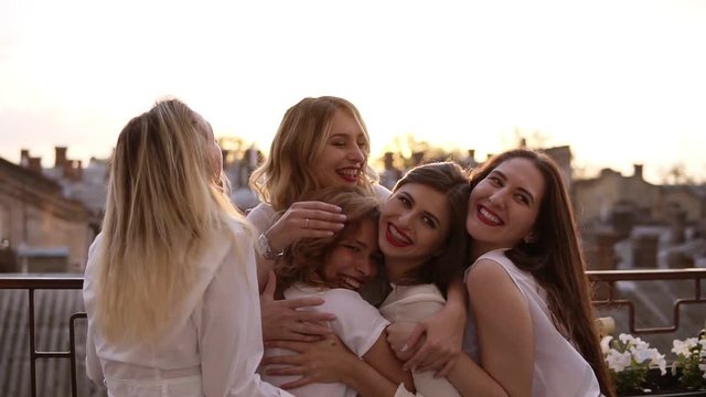 Attractive caucasian girls are hugging standing outside on a terrace or balcony. Six beautiful young women in white shirts and with red lipstick. Hen party. Evening dusk. Love and friendship