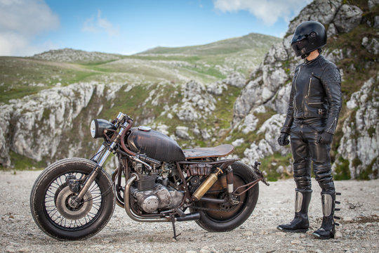 Biker with black leather suit and mask stay next his custom special rat motorbike.  Desolated mountains landscape in the background. Post apocalyptic concept