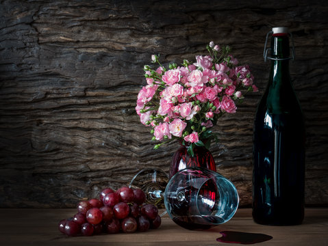 Still life visual art of grapes, a falled glass of wine, a bottle of wine and pink roses in a red vase on wooden slab with wooden back drop
