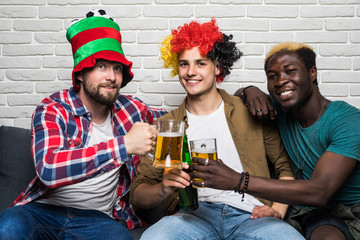 Three happy men sitting on couch and watching sport on TV and cheers beer