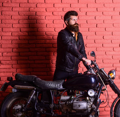 Hipster, brutal biker on serious face in leather jacket gets on motorcycle. Start of journey concept. Man with beard, biker in leather jacket near motor bike in garage, brick wall background.