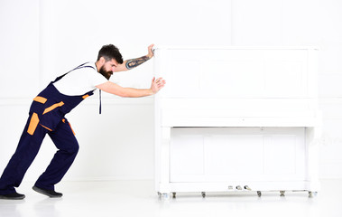 Man with beard and mustache, worker in overalls pushes piano, white background. Loader moves piano...