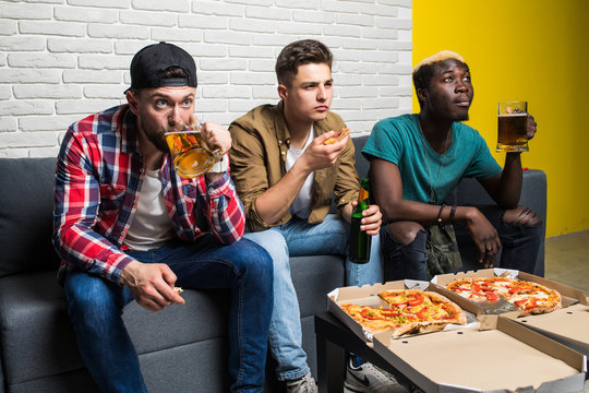 Group Of Three Male Friends Watching A Soccer Game On TV While Drinking Beer And Eating Pizza