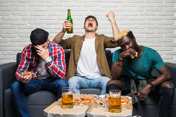 Three young mixed race men sitting on sofa drink beer and eat pizza support different team and with different emotion of lose and win at home