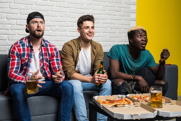 Attractive guys are drinking beer, eating pizza, talking and smiling while resting on sofa at home