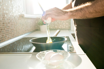 Close-up of unrecognizable man in apron with hairy arms pouring egg into cooking pan while cooking...