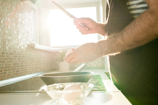 Close-up of unrecognizable man cooking fried eggs on induction cooktop and cracking egg with knife in bright sunlight from window
