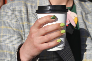 Fototapeta premium Beautiful female hand with manicure holds cardboard coffee cup. Tender yellow nail polish and green cat eye design.