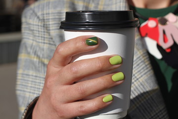 Beautiful female hand with manicure holds cardboard coffee cup. Tender yellow nail polish and green...