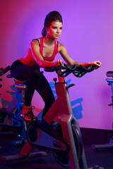 Fototapeta na wymiar Gorgeous brunette woman with long straight hair pulled back in tail, wearing fitness top and leggings, doing cardio exercise on stationary bicycle in pink neon light during workout in sports club.
