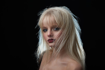 Model with blonde dishevelled hair and opened shoulders looking at camera posing on black studio background. Girl having blue eyes, plump lips covered with red lipstick, wearing evening make up.