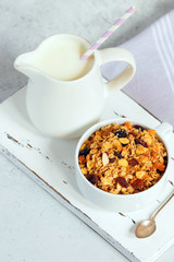 Cup of muesli with tropical fruits and white milk jug on wooden board. Concept of healthy eating in pure tones.