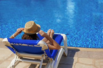 Fototapeta na wymiar Woman in a straw hat relaxing on a deck-chair near a luxurious summer pool in hotel, concept time to travel