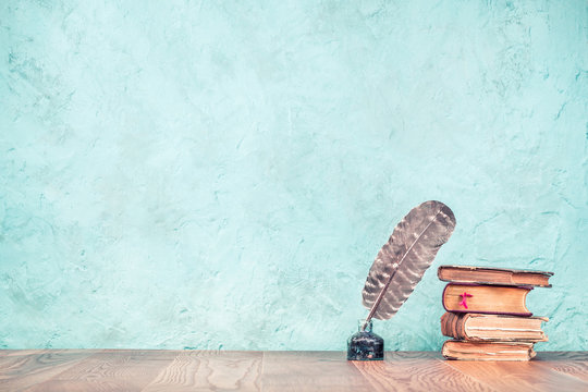 Quill ink pen with inkwell and aged books on wooden desk front aquamarine concrete wall background. Vintage old style filtered photography