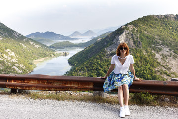 Fototapeta na wymiar Woman with shot ginger hair is sitting on the bumper on the mountains and river background. active lifestyle, summer holiday concept. backview