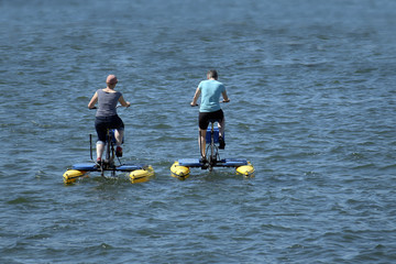 man and woman from behind ride with floating pedal bicycle boats across the blue lake, holiday and...