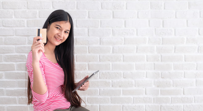 Asian woman in casual cloth holding credit card and using to online shopping with tablet at white brick wall.Digital technology lifestyle.Banner with copy space for display text or design.