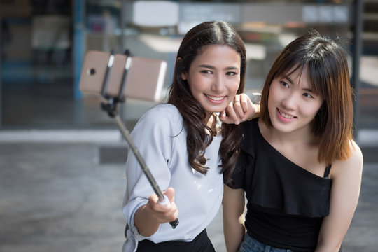 happy woman best friend taking selfie photograph together; smiling asian girl with her bestie using selfie stick to take hobby, leisure, casual selfie photo in urban environment; asian 20s woman model