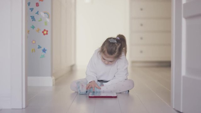 Casual little girl sitting on floor in doorway of apartment while using tablet and looking interested in game. 