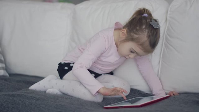 Charming little girl sitting on cozy big couch at home and browsing tablet looking absorbed into gadget usage. 