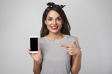 Fototapeta premium Isolated studio shot of adorable ecstatic young lady with dark hair having excited happy expression, advertising modern smart phone, pointing at copyspace screen on generic electronic gadget
