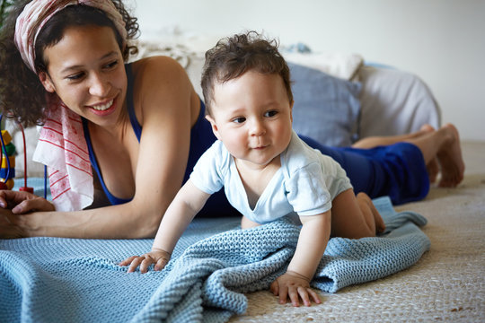 Indoor shot of cute baby boy crawling on floor in living room, studying and touching various objects, having curious playful expression, his young mother lying behind him and smiling happily