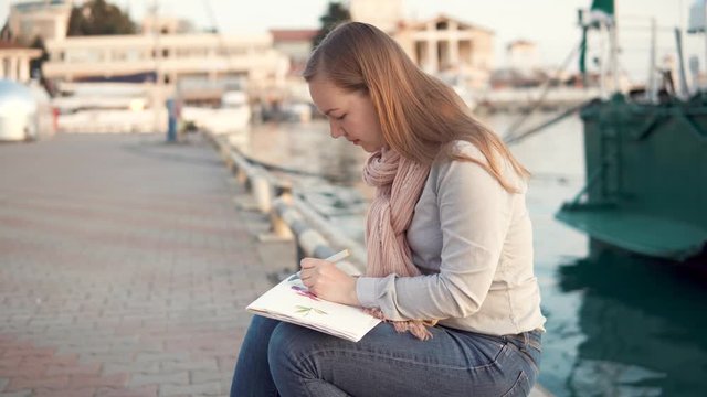Side view shot of a beautiful inspired artist drawing picture sitting at the bay in the evening. Doing coloring in sketchbook using markers.
