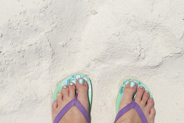 Fototapeta premium Girl feet in blue slippers on sand beach texture. Tropical seashore. Seaside banner template with text place.