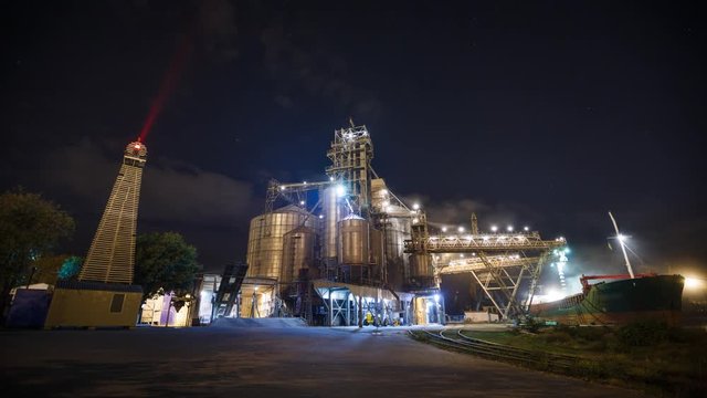 Night timelapse of loading grain crops on bulk freighter ship via trunk to open cargo holds at silo terminal in seaport from trucks. Cereals bulk transshipment. Transportation agricultural products.